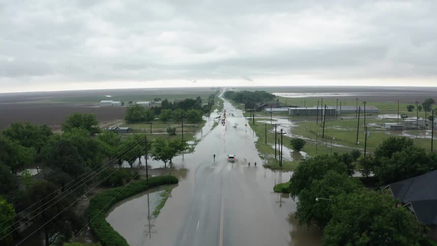 A car leaves a wake as it travels on a flooded road while small children play in the splashing water