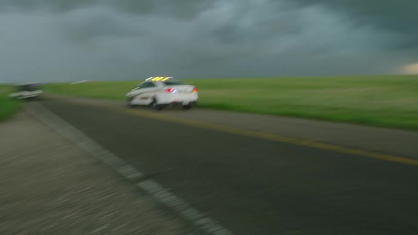 A police car with sirens on rushes by a middle aged man on the way to the scene of tornado destruction after a severe storm caused damage to a small town