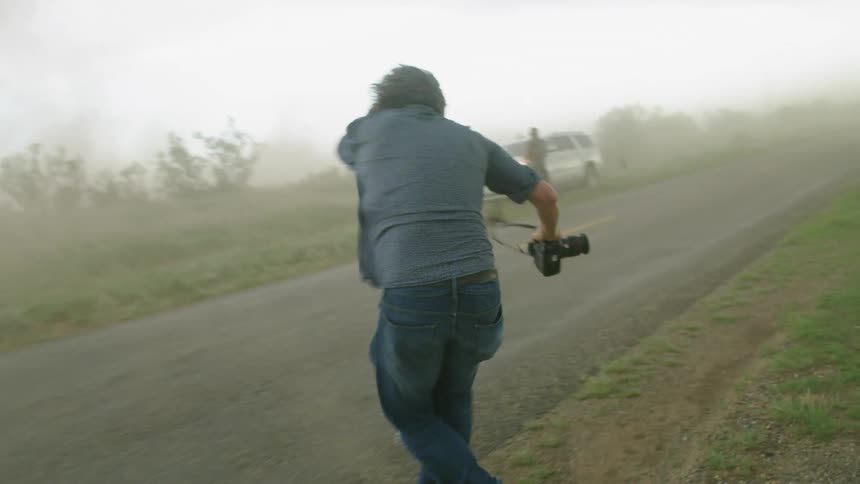 A middle aged man runs across a rural road as severe thunderstorm winds blow fierce sand and dust horizontally across the frame