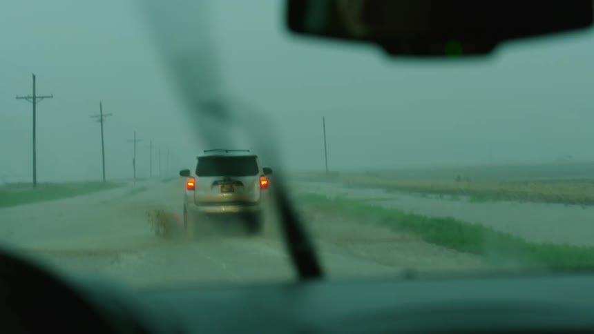 Windshield wipers seen from the inside of a a moving car that is driving through a large flooded highway following another vehicle with its lights flashing
