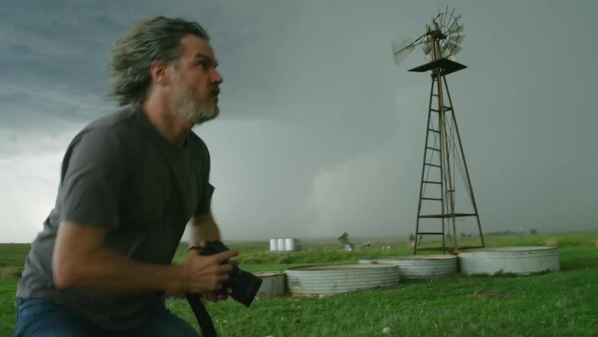 A middle aged male photographer man kneeling in a grassland raises his camera to photograph a tornado before standing and running away in this high energy high intensity shot