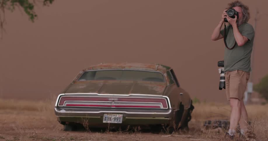 A middle age photographer takes photos of a dust storm haboob as the junkyard and abandoned car are overwhelmed by blowing sand