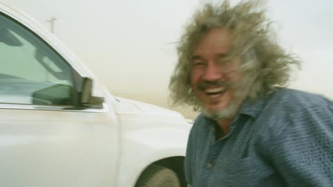 A Middle Aged Man laughs at camera as hurricane force winds blow sand, dust and his hair wildly in the wind while stormchasing