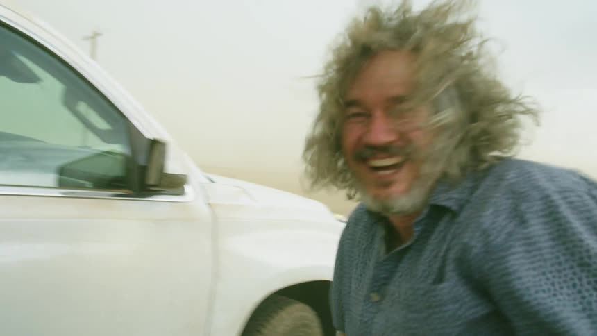 A Middle Aged Man laughs at camera as hurricane force winds blow sand, dust and his hair wildly in the wind while stormchasing