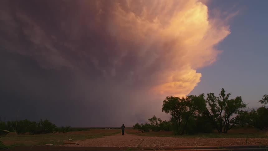 Real time shot of a man walking down a rural dirt road with a huge anvil shaped thunderstorm illuminated by glowing orange, purple and pink sunset light while large lightning flashes in the supercell storm cell