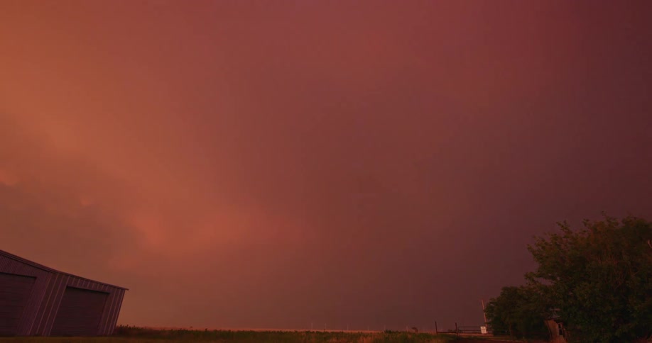 Anvil crawler lightning strikes left and down across a hazy orange red sky over a flat green land near a steel building