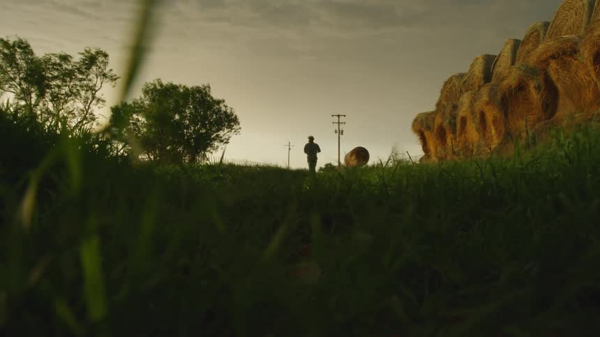 Wind whips grass in the foreground as a man walks past a large stack of hay bales with severe thunderstorm clouds above him