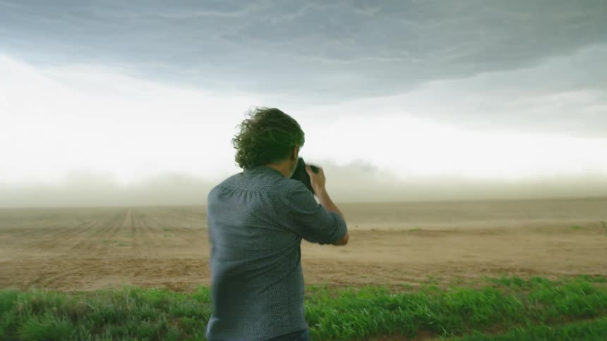 A silver and brown haired adventurous stormchaser man photographs severe supercell thunderstorm winds that blow topo soil away from the corn fields