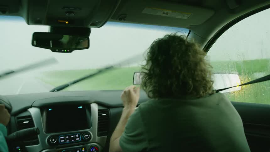 Man sitting in the passenger seat of a moving car filming a supercell thunderstorm while hail pounds the vehicle on the interstate