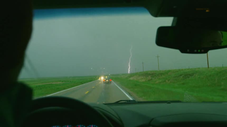 Inside of a vehicle with its windshield wiper on driving down a road towards a severe storm with strong lightning bolts