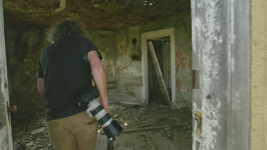 A photographer carefully walks into an abandoned rural farm house to explore the surroundings and document the scene