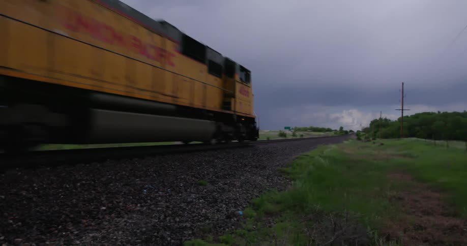 A freight train locomotive passes as the camera whip pans to follow the locomotive and freight cars with dark stormy clouds  as it passes by