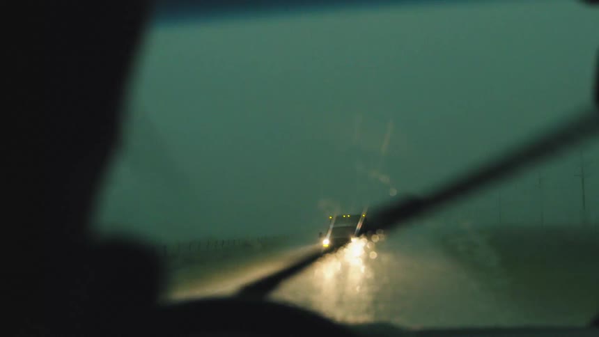An oncoming cards headlights illuminate the interior of the vehicle and the windshield coated with heavy thunderstorm rain in slow motion