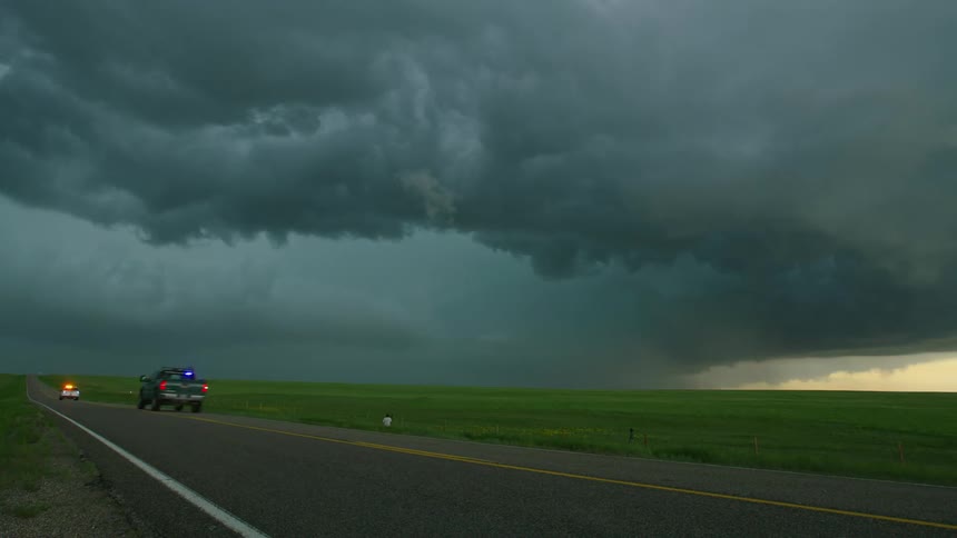 Emergency vehicles drive down a rural highway approaching a town after a tornado destroyed several houses, the severe storm can be seen in the background
