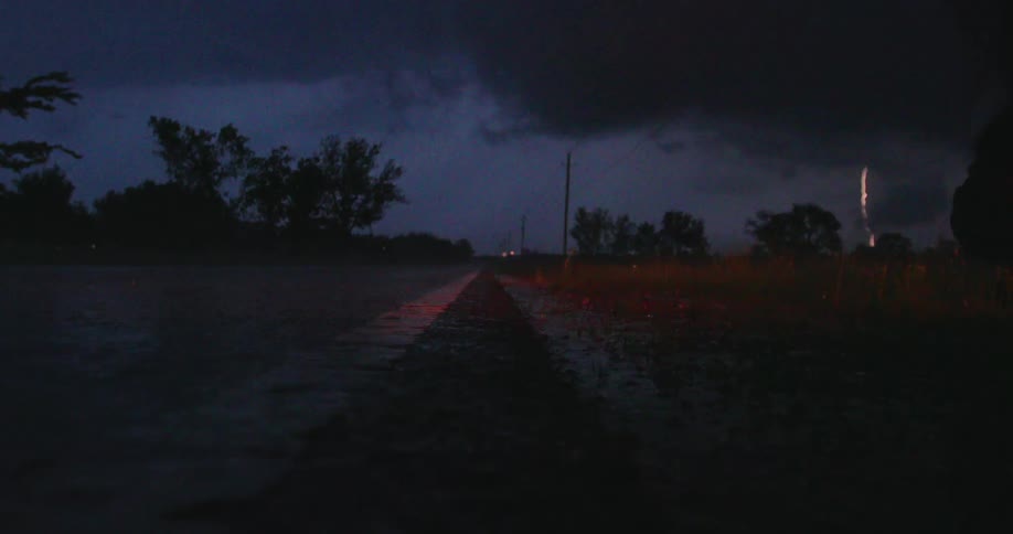 Real time shot looking down the painted line of a wet roadway at night with storm clouds swirling and intense lightning bolts on the right side of the frame