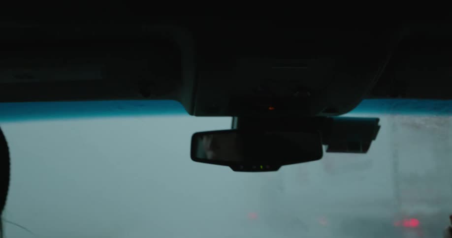 A woman drives through heavy rainfall and large hail stones as she passes semi trucks with their hazard lights on and we see her eyes in the rear view mirror
