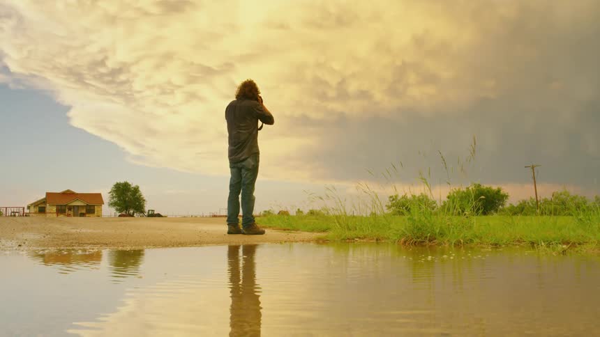 A man takes photos of a massive supercell thunderstorm at sunset  while golden light illuminates the mammatus clouds and puddle foreground