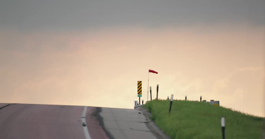 A red cabin semi truck drives down a hill as the wind blows a windsock horizontal and blows through the tall grass alongside highway