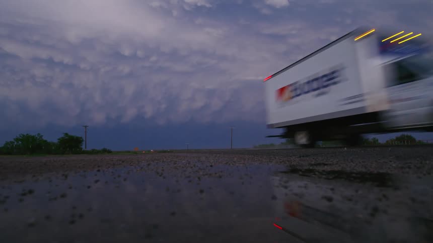 The camera is low on the rain soaked ground looking at a huge thunderstorm after the sun has set and lightning strikes and a large truck with headlights drives by