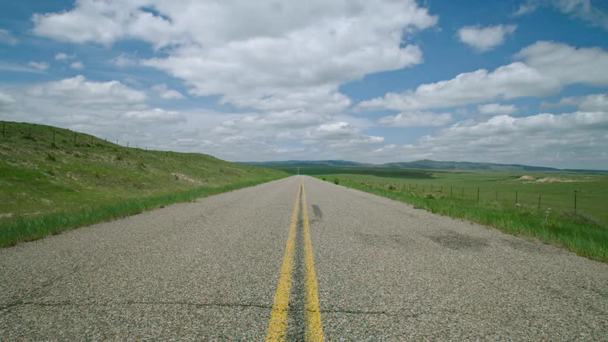 Time lapse of center framing of a rural road and green rolling hills as clouds pass by a blue sky