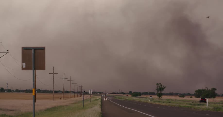 Real time shot looking down a highway system with car headlights and birds flying in the foreground and massive plumes of blowing dust and sand in the background