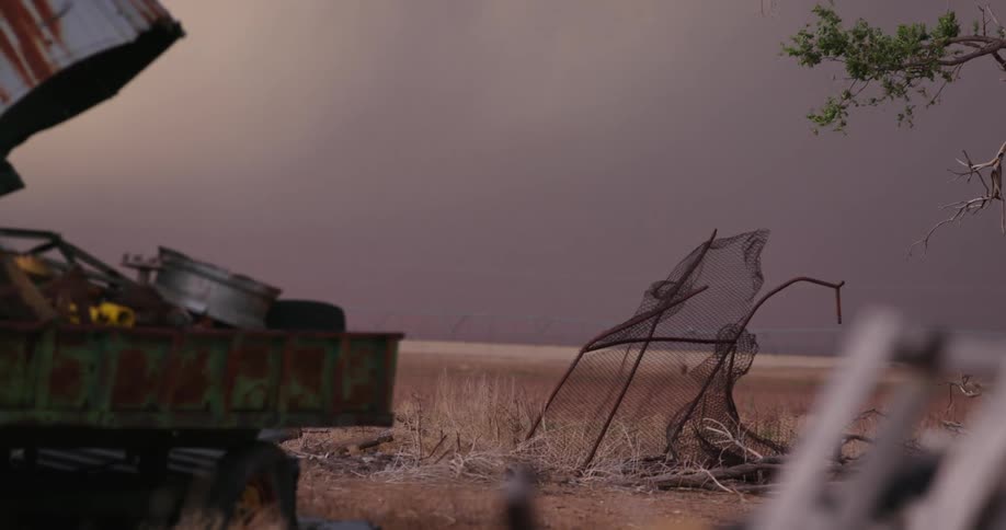 Dark and stormy skies are over an abandoned outdoor space with a destroyed fence and weeds in the ground as dust storm approaches