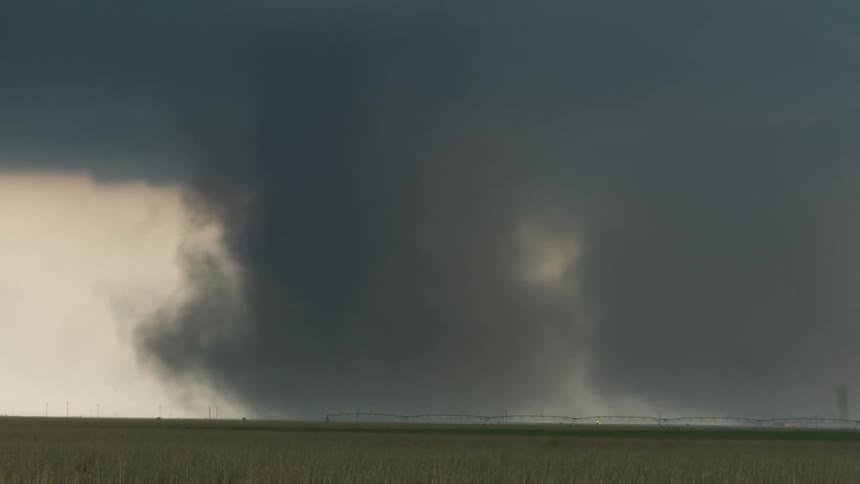 An enormous and menacing tornado spins above a wide open grassy plain with center pivot sprinklers and passing cars for scale in the foreground