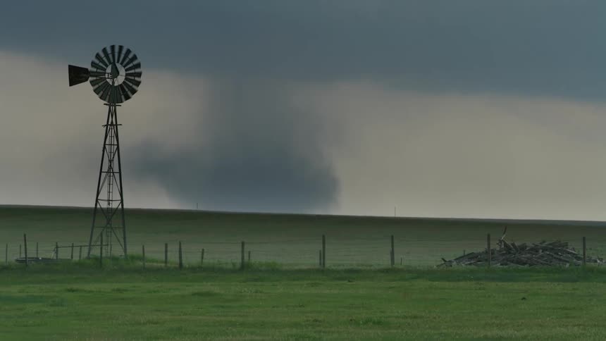 Real time shot of a tornado on the ground behind an open field with a barbed wire fence a pile of wood and a weather vein