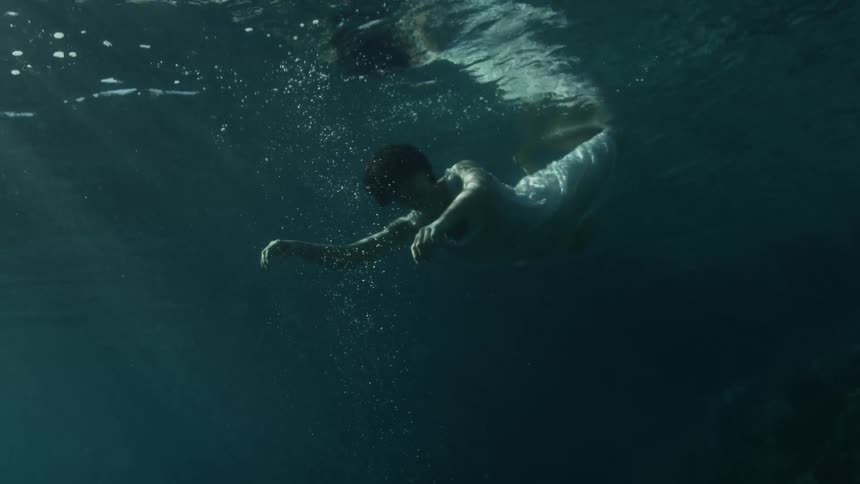 A woman in a white gown floats lifelessly under water and gradually floats to the surface