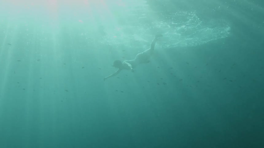 Sun light shining down underwater in the ocean as a woman swims gently to the surface of the water