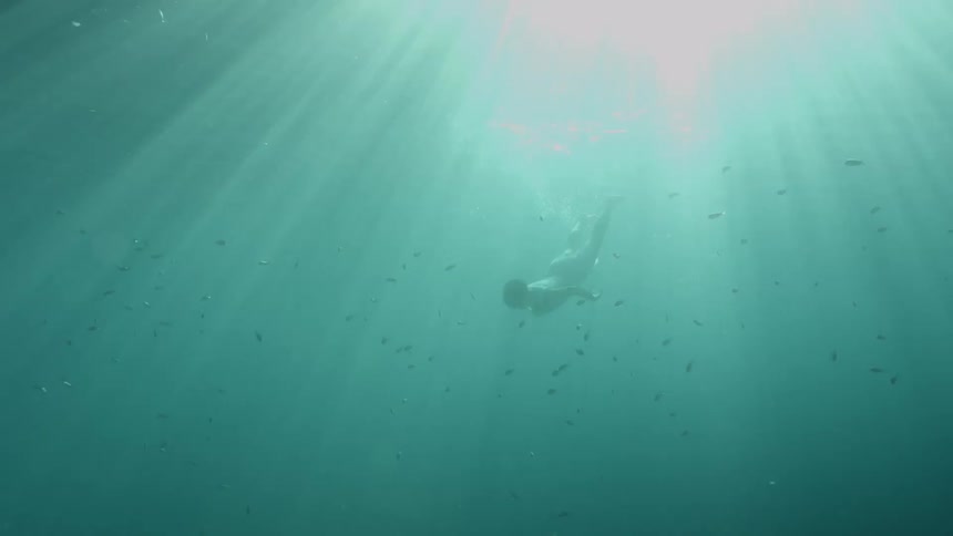 Sunlight shines down on a woman as she swims underwater surrounded by dozens of fish