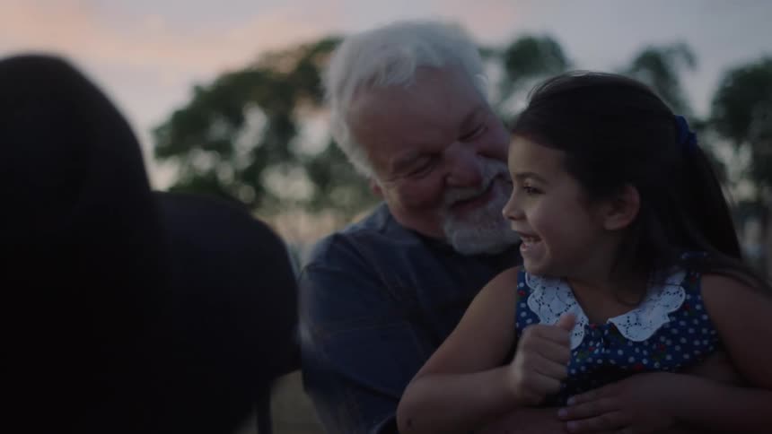 A man holds his granddaughter in his lap as she smiles while petting a horse