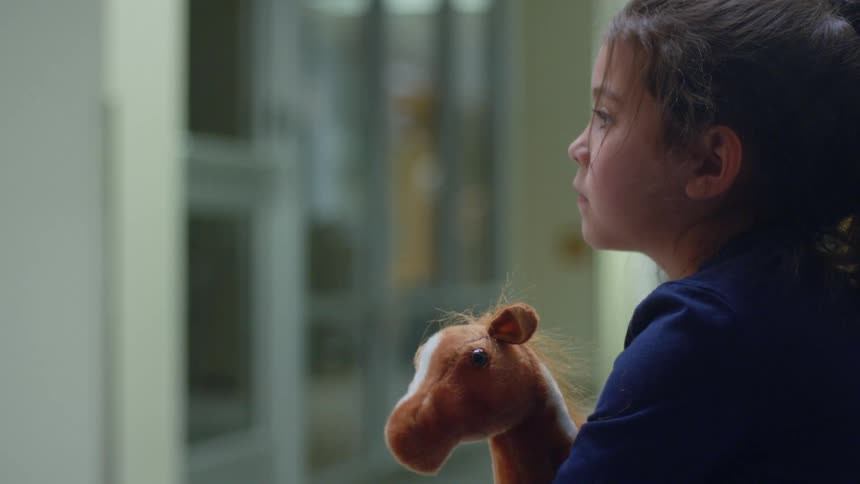 A toddler clutches a stuffed giraffe as she waits in a hallway in a building