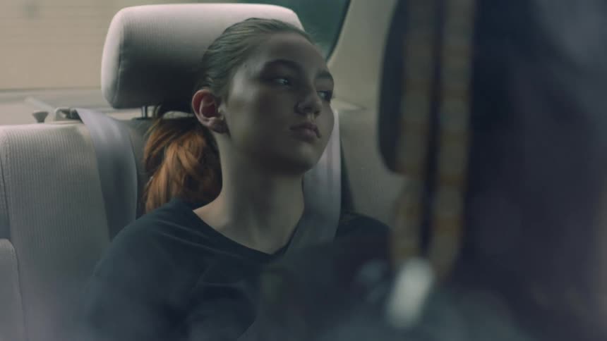A teenage girl sits in the backseat of a car looking bored
