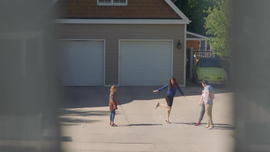 A dad hugs his daughter as they watch mom and sister hopscotch in the driveway