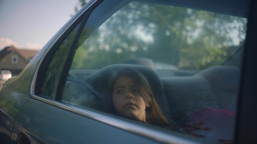 A young girl sits in the backseat of her familys parked vehicle and looks out the window