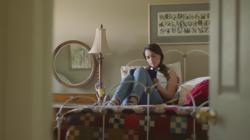 A young woman sitting on a bed with her back resting against a pillow uses a pencil to write on a binder
