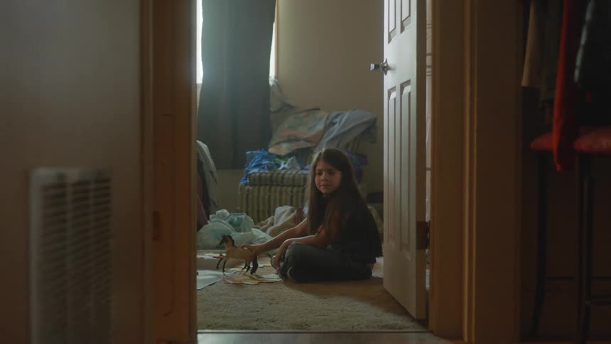 A girl with long dark hair sits on the floor of her bedroom and plays with a plastic toy horse