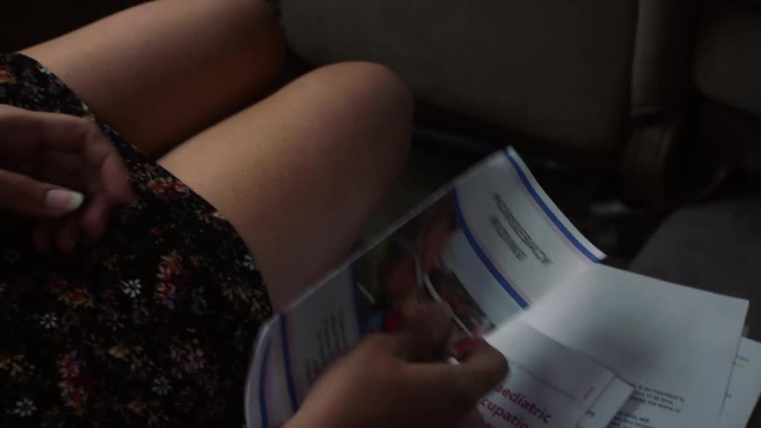 A woman sitting in the backseat of a moving vehicle lifts a pamphlet from the seat beside her and holds it in her lap