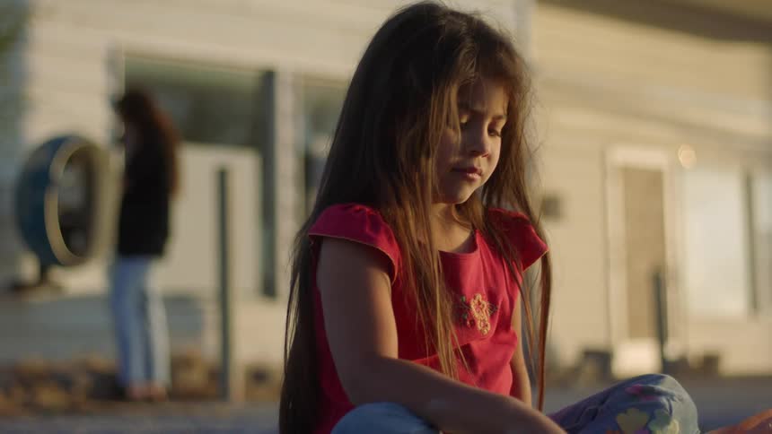 A young girl sits an calmly plays as another older female talks on a phone in the distance