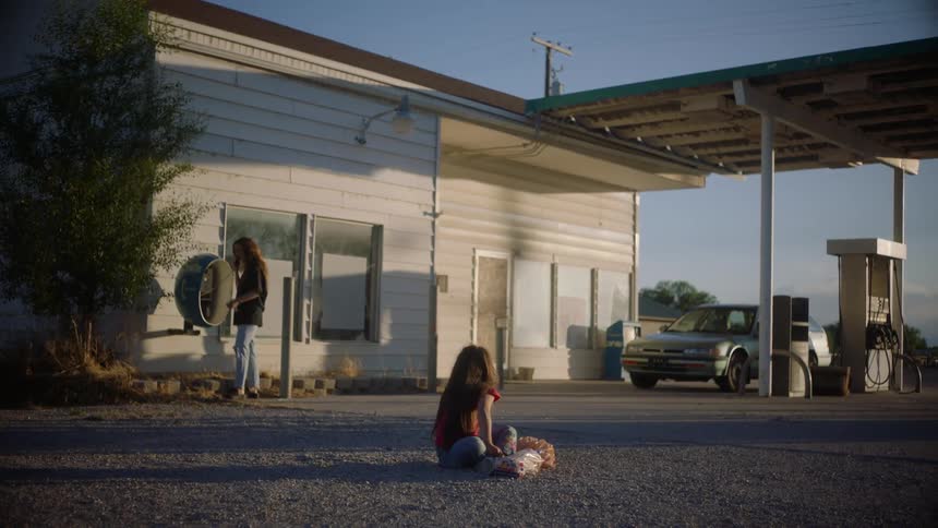 A woman uses a phone booth at a gas station in a rural area as her daughter sits on the ground with her doll