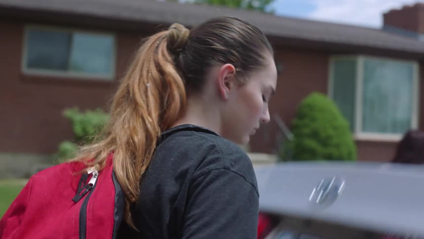 A woman pulls a suitcase out of the trunk of a vehicle while wearing a backpack