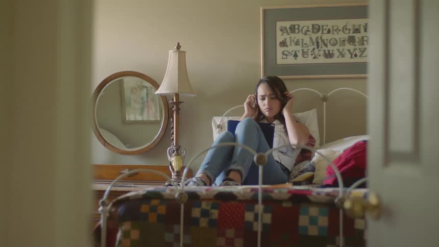 A teenage female reclines on her bed with a notebook as she frowns before putting on her headphones