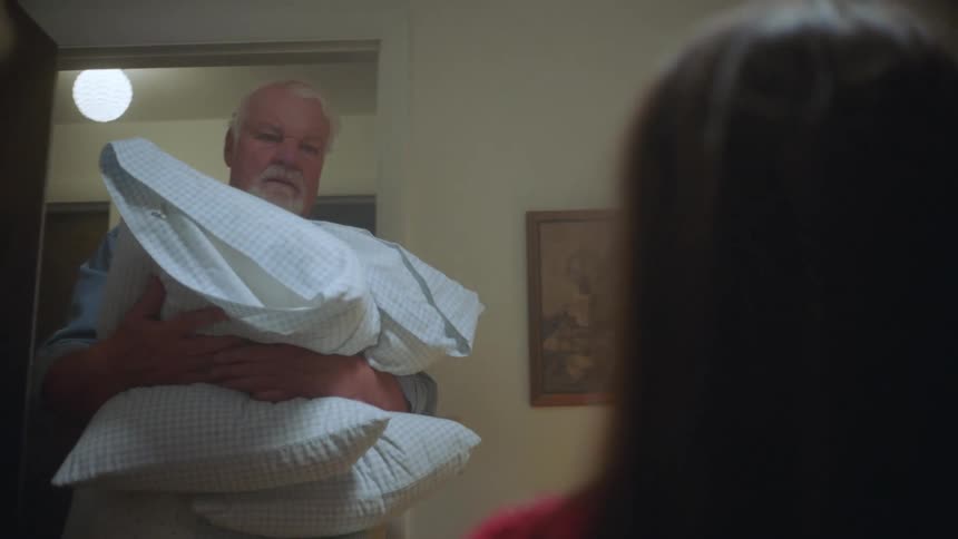 A young girl sits in a room as an elderly man enters carrying pillows