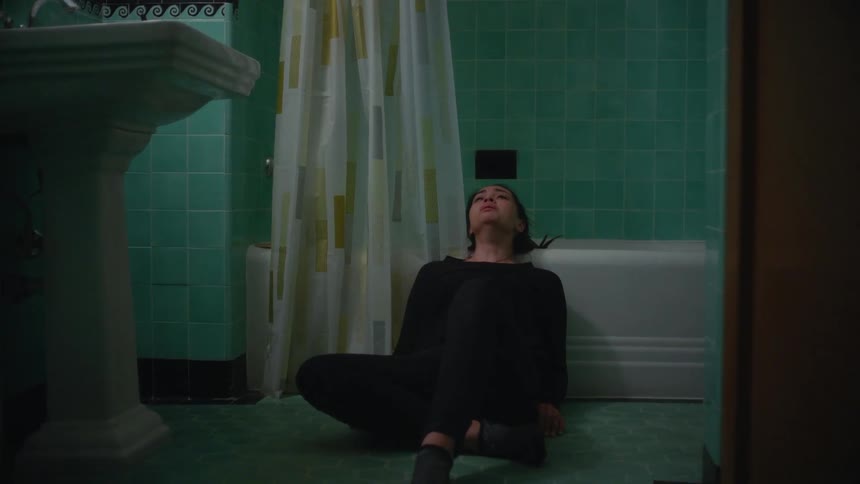A crying woman in black rests her head on the bathroom sink and collapses backwards onto the bathroom floor and edge of the bathtub