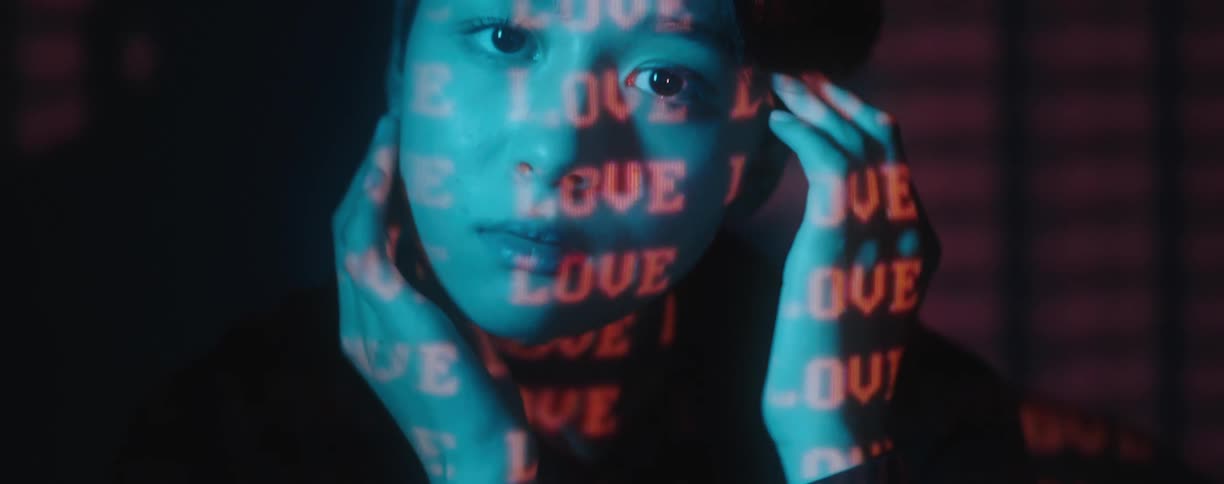 Young woman rubs face and and neck with hands while the word love is projected in red on her face neck and the wall behind her