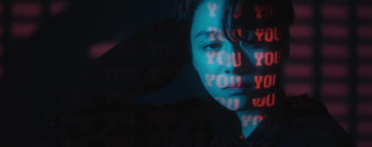 A young asian man sits in front of a dark wall running a hand through his hair while looking forward and the word you projects in a pattern onto his face