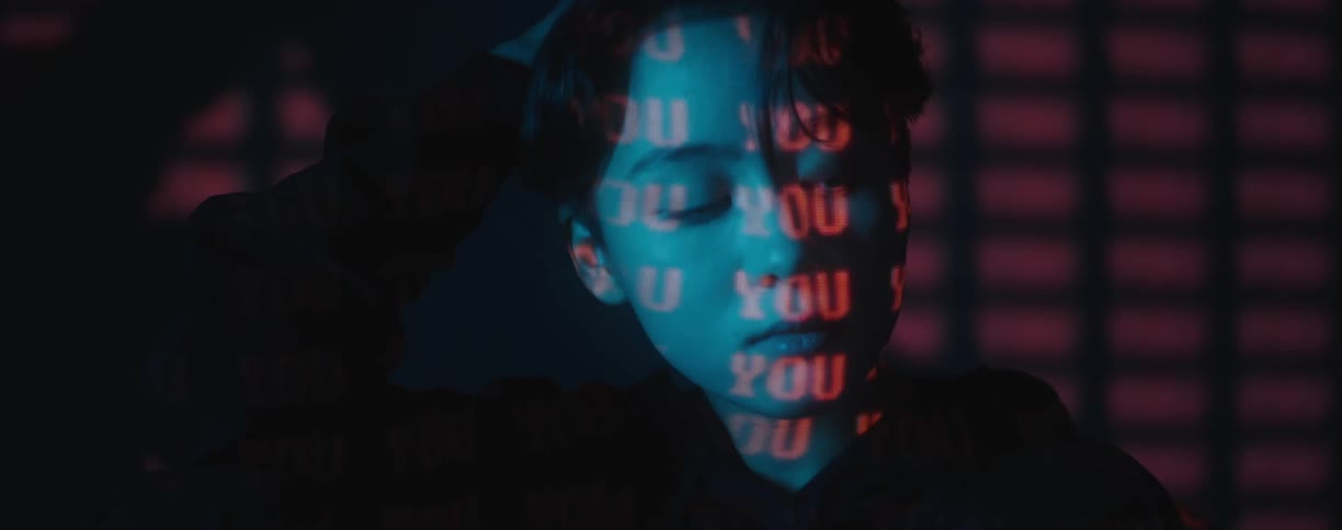 A woman with short hair sits in a dark room with red words projected onto her