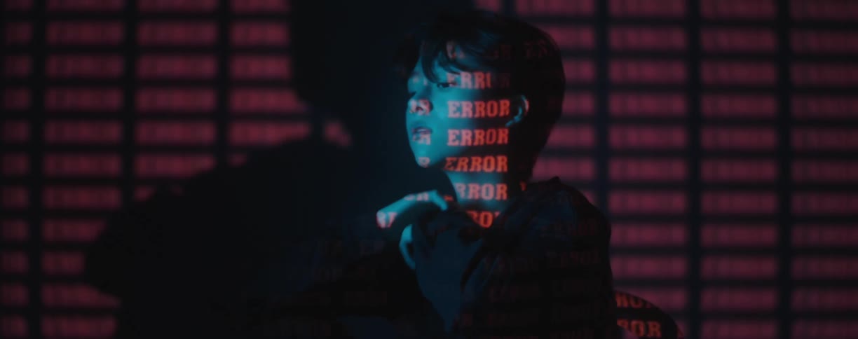 A bored woman with short hair sits at a table in a dark room with red words projected onto her
