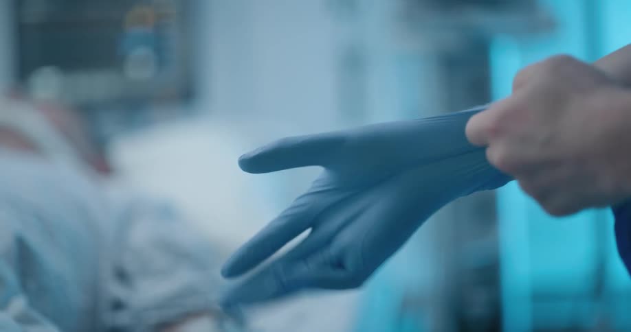 A person places blue gloves on their hands as they stand in a hospital room with a patient in the bed